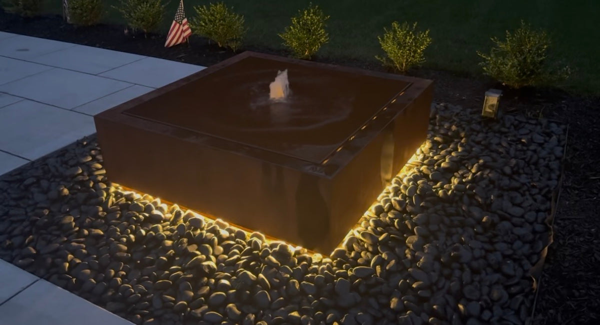 Square Corten steel fountain at dusk with warm LED lighting under its base, surrounded by black river stones, customer photo