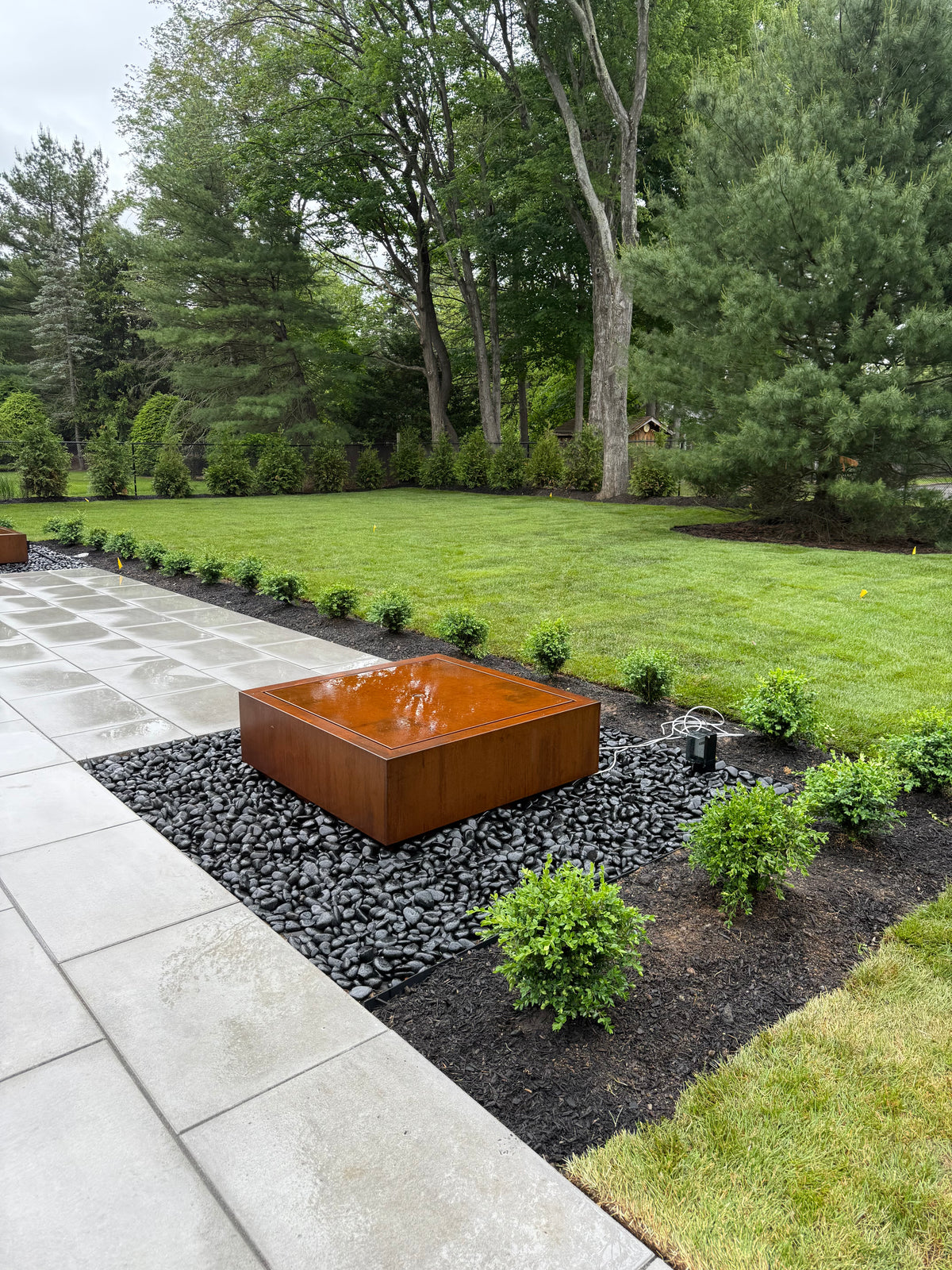 Square Corten steel fountain with a water-covered top in a black river stone bed beside a paver patio, customer photo