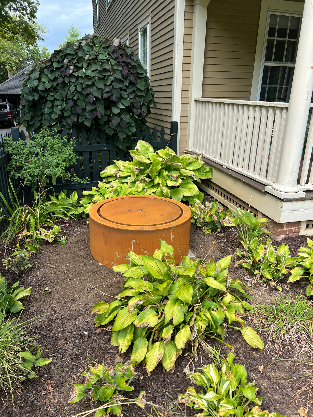 Round Corten steel fountain installed beside a covered porch in a garden of hostas, customer photo