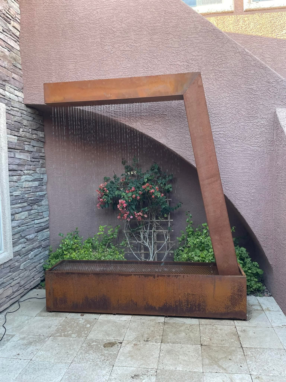 Custom Corten steel rain curtain fountain with an angled frame above a Corten basin, in front of a bougainvillea and a stucco wall, customer photo