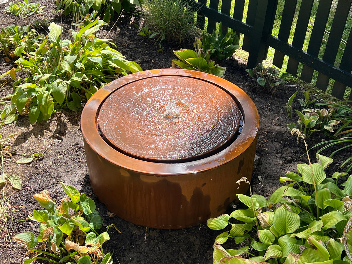 Corten steel round fountain running in a garden bed surrounded by hostas, customer photo