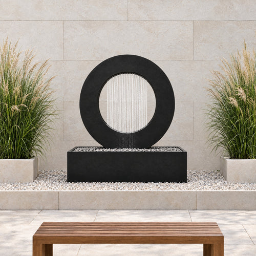 Black steel Halo rain curtain fountain in a gravel bed against a limestone wall with ornamental grasses