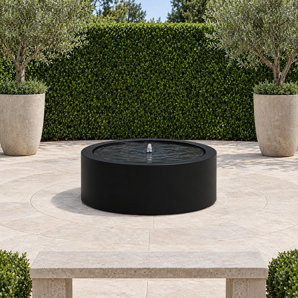 Black steel Mesa Circular fountain in a limestone courtyard in front of a clipped hedge and two olive trees