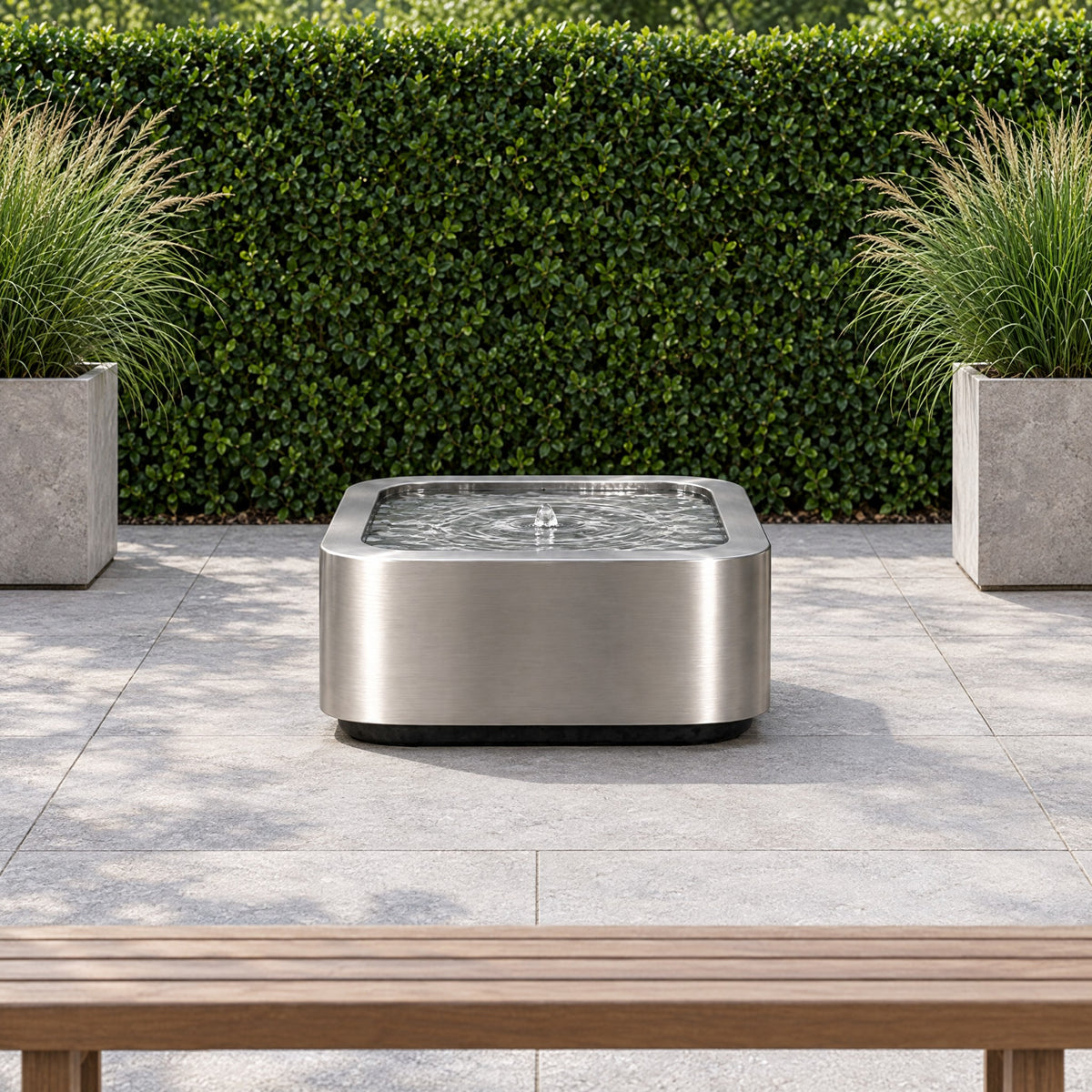 Stainless steel Mesa Soft Square fountain in a garden courtyard with a hedge wall and grass planters